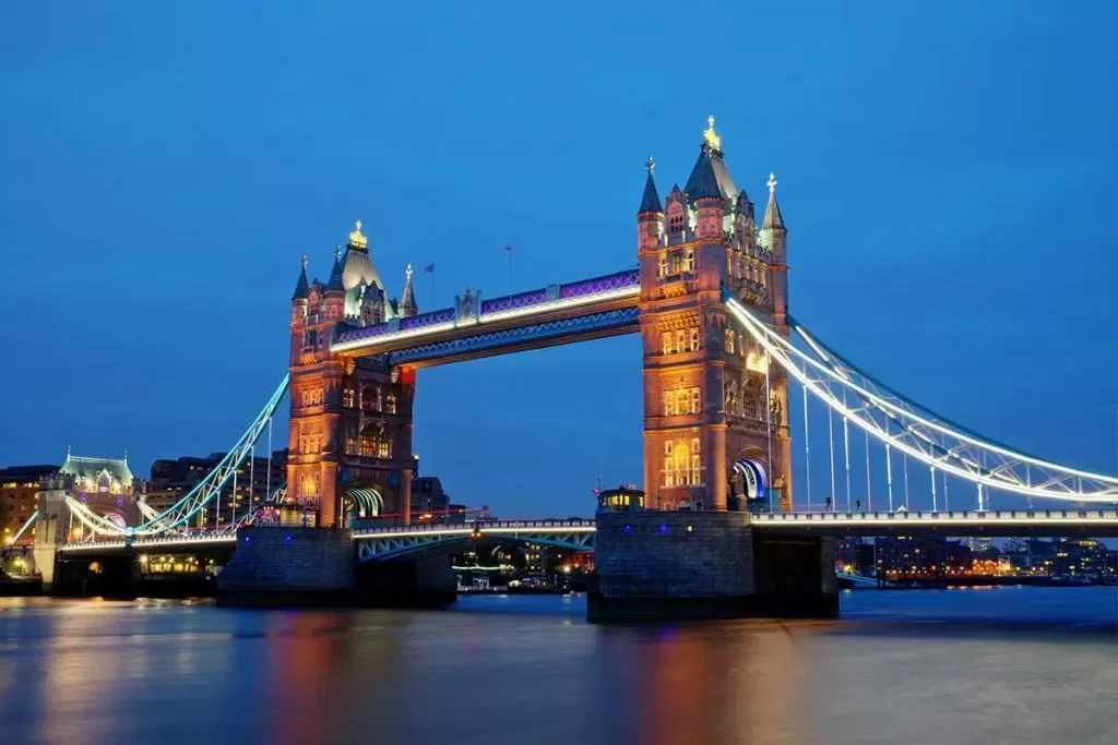 Nighttime view of the Tower Bridge in London, illuminated with colorful lights, over the River Thames.