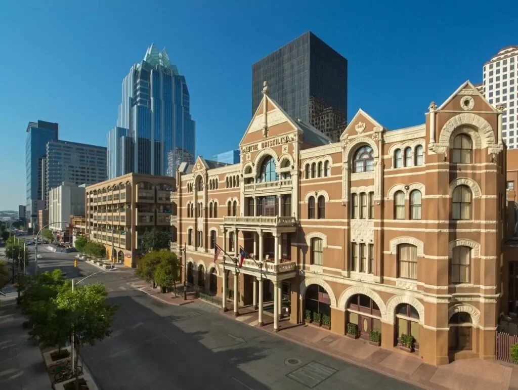 Historical brick building with arched windows and a balcony, surrounded by modern skyscrapers in an urban setting.