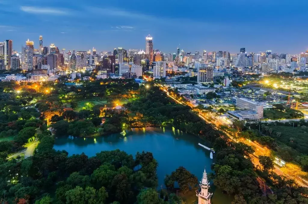 Nighttime aerial view of a city skyline with illuminated buildings and a park with a lake in the foreground.
