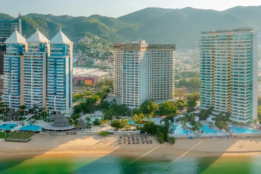 View of a beach resort with tall white and blue buildings, pool areas, and a sandy beach front with lounge chairs, set against a backdrop of green hills and a cityscape.