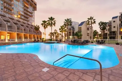 Empty outdoor swimming pool with clear blue water, surrounded by apartment buildings and palm trees at sunset.