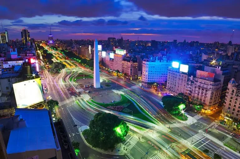Nighttime city view with illuminated streets, buildings, and a tall obelisk monument in the center of a roundabout, with vibrant light trails from moving vehicles.