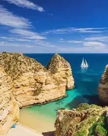 Sandy beach with large cliffs and a sailboat on the ocean in the background under a partly cloudy sky.