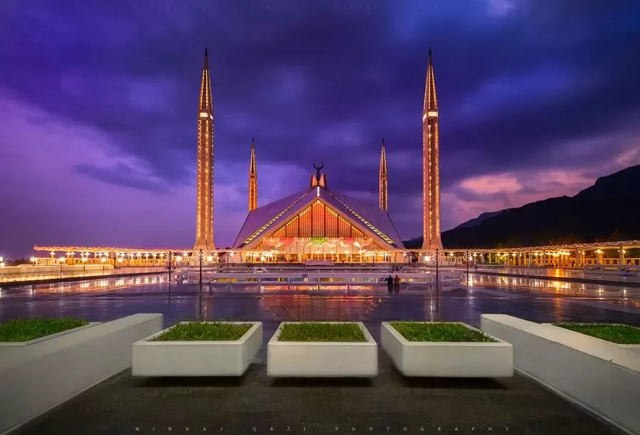 The image shows the Faisal Mosque in Islamabad, Pakistan, illuminated at night with a cloudy sky in the background.