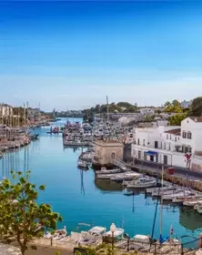 A scenic harbor with boats docked along the pier, white buildings lining the waterfront, and a clear blue sky overhead.