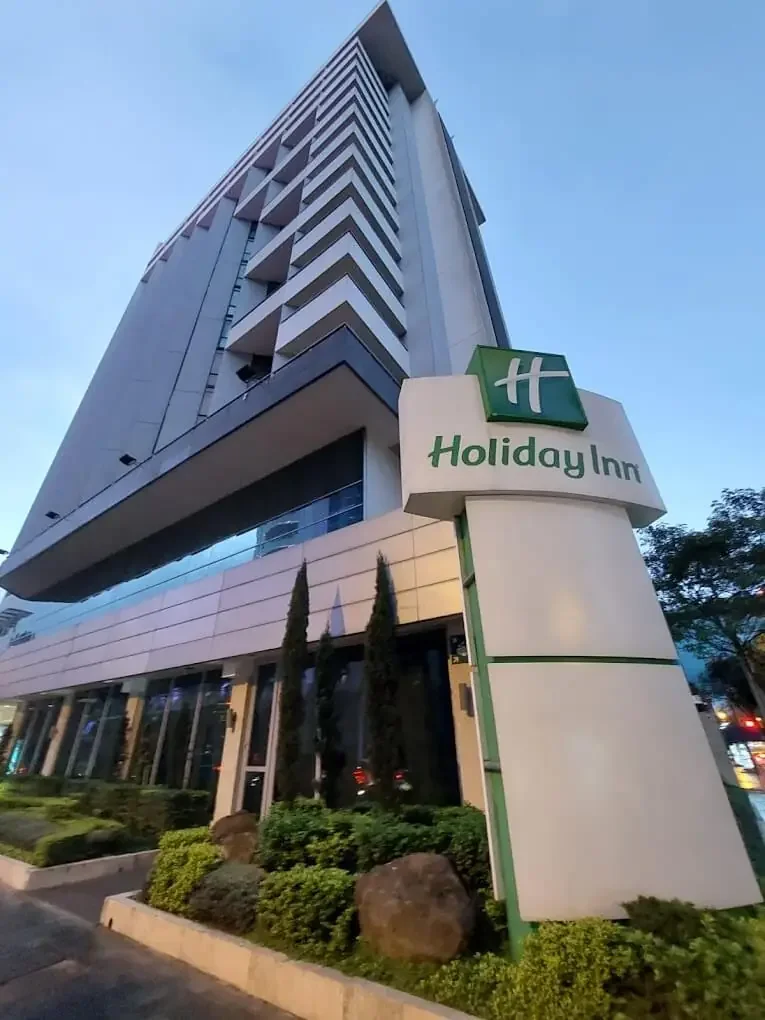 A tall, modern hotel building with the Holiday Inn sign near the entrance, surrounded by landscaped plants and rocks.