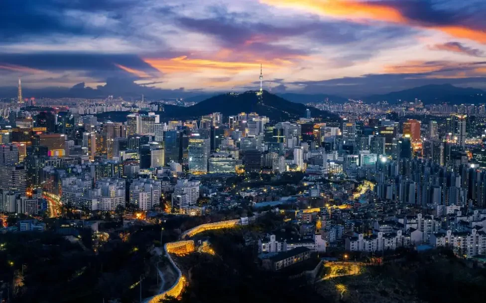 Nighttime cityscape of Seoul, South Korea, with high-rise buildings, Namsan Seoul Tower on a hill, and a colorful sunset sky.