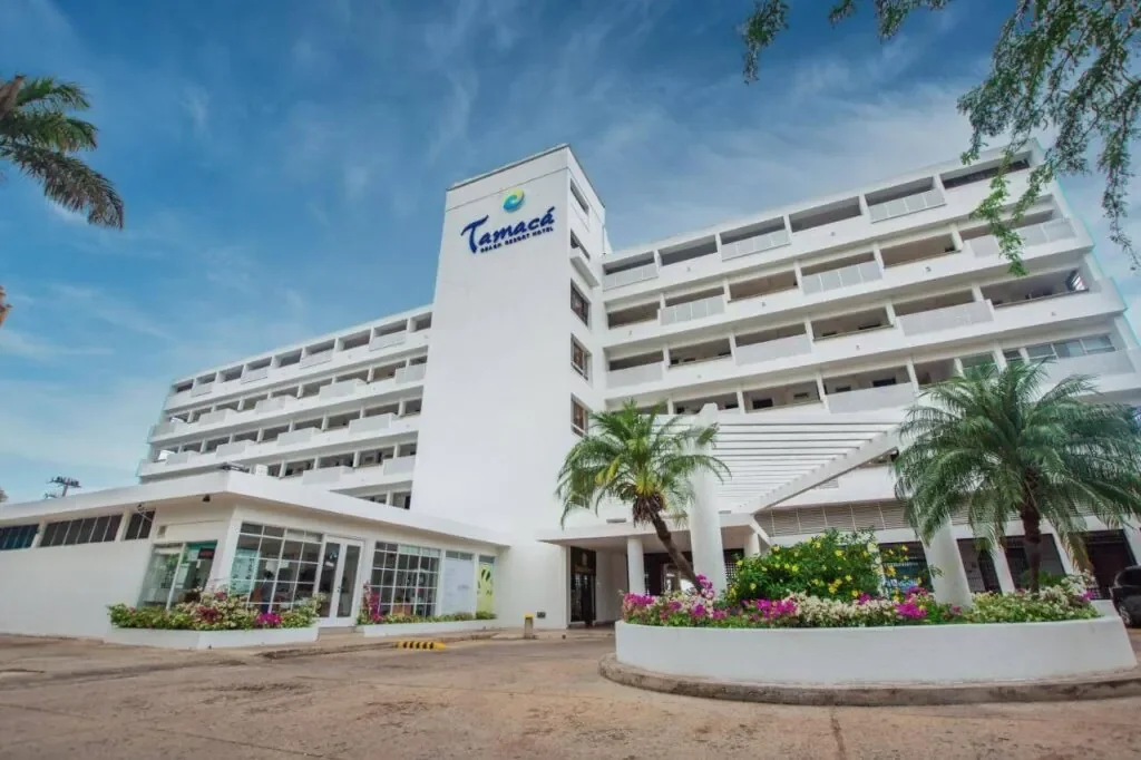 Hotel Tamaca in Barranquilla, Colombia, with a modern white facade, palm trees, and colorful flower beds in the foreground.