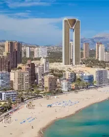 A city skyline with a distinctive tall building that has an arch-shaped top, situated along a beach with people and umbrellas, under a clear blue sky.