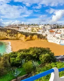 A scenic coastal view with a sandy beach, green foliage, and colorful buildings on a hill under a partly cloudy sky.