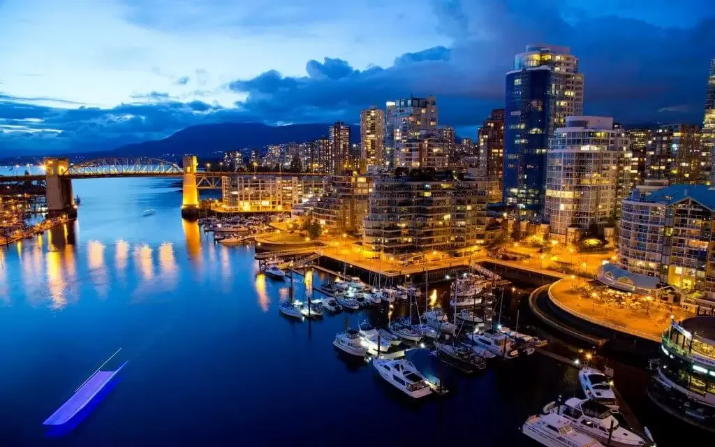 Night view of a city waterfront with high-rise buildings illuminated, boats docked in the marina, and a bridge over the water, with mountains in the background.