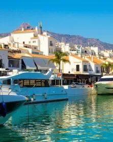 A marina with boats docked on calm water, beachfront buildings with balconies, palm trees, and a mountainous background under a blue sky.