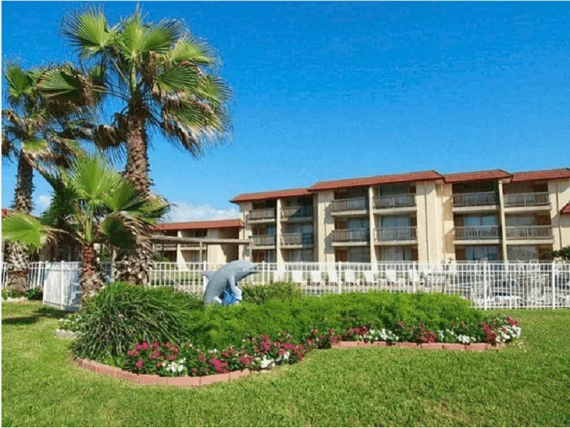 A landscaped garden with palm trees and colorful flowers in front of a multi-story resort building under a clear blue sky.