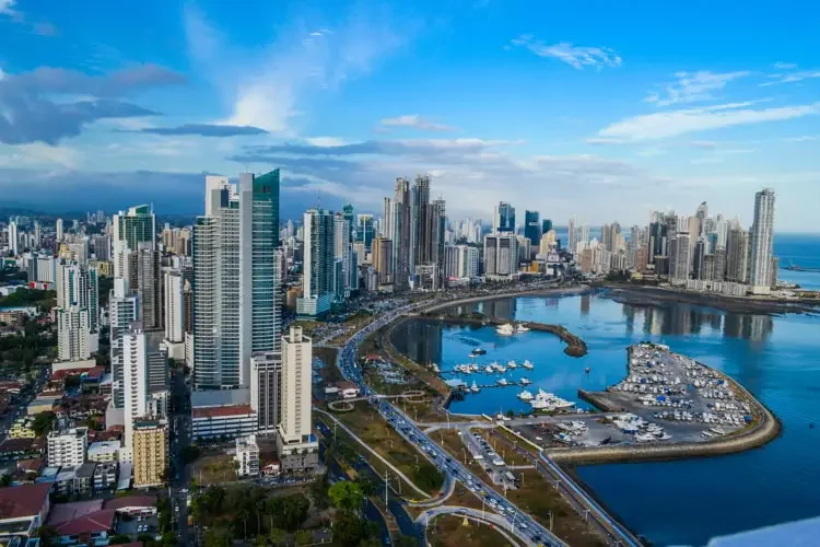 Aerial view of a modern city skyline with high-rise buildings, a harbor with boats, and a curved road near the water under a partly cloudy sky.