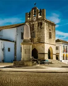 Historic stone church with a bell tower, situated in a town square with a statue of a religious figure in front, and colorful colonial buildings nearby under a blue sky.