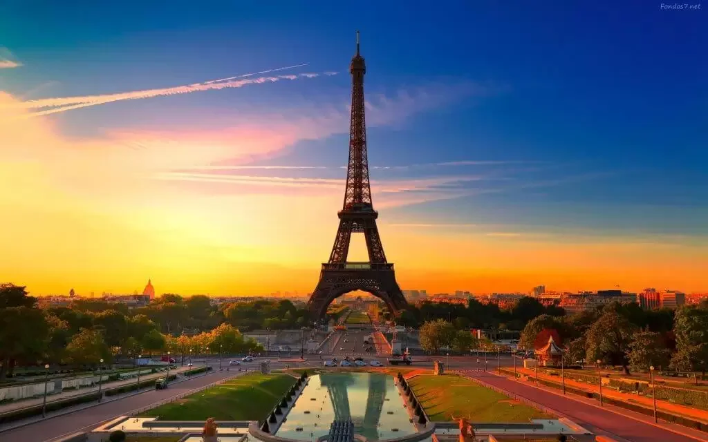 Sunset view of the Eiffel Tower in Paris, France, with reflections in a water feature in the foreground and trees lining the scene.
