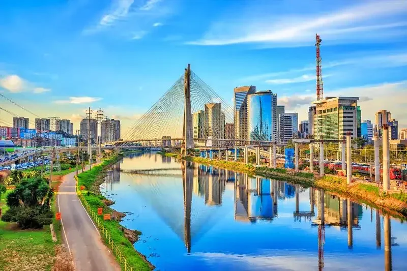 A city skyline with modern skyscrapers and a cable-stayed bridge over a river, under a blue sky with scattered clouds.