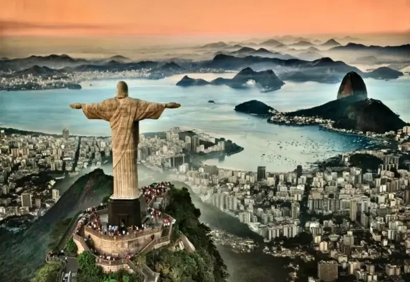 A large statue of Jesus Christ with arms outstretched overlooking Rio de Janeiro, Brazil, with city buildings, Guanabara Bay, and mountains in the background at sunset.