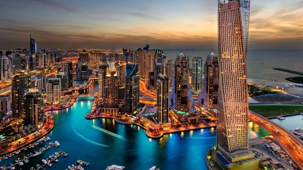 Aerial view of Dubai Marina at sunset with tall skyscrapers, a winding canal, and boats docked along the water.