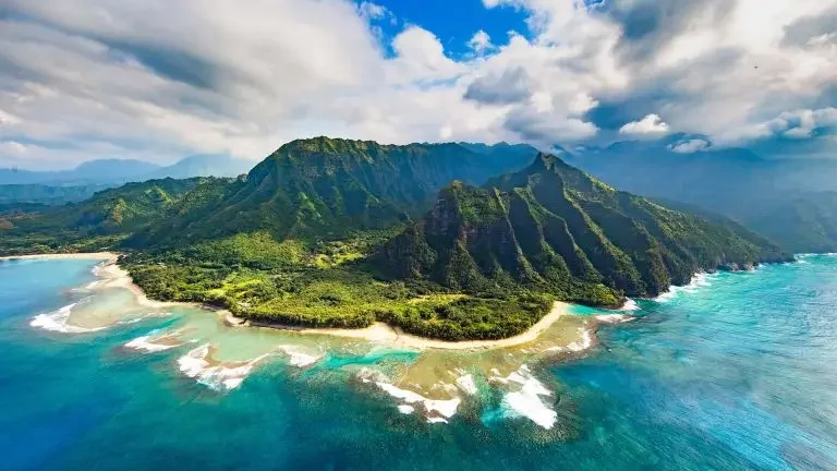 Aerial view of a lush green island with mountain terrain, surrounded by vibrant blue ocean waters and white sandy beaches under a cloudy sky.