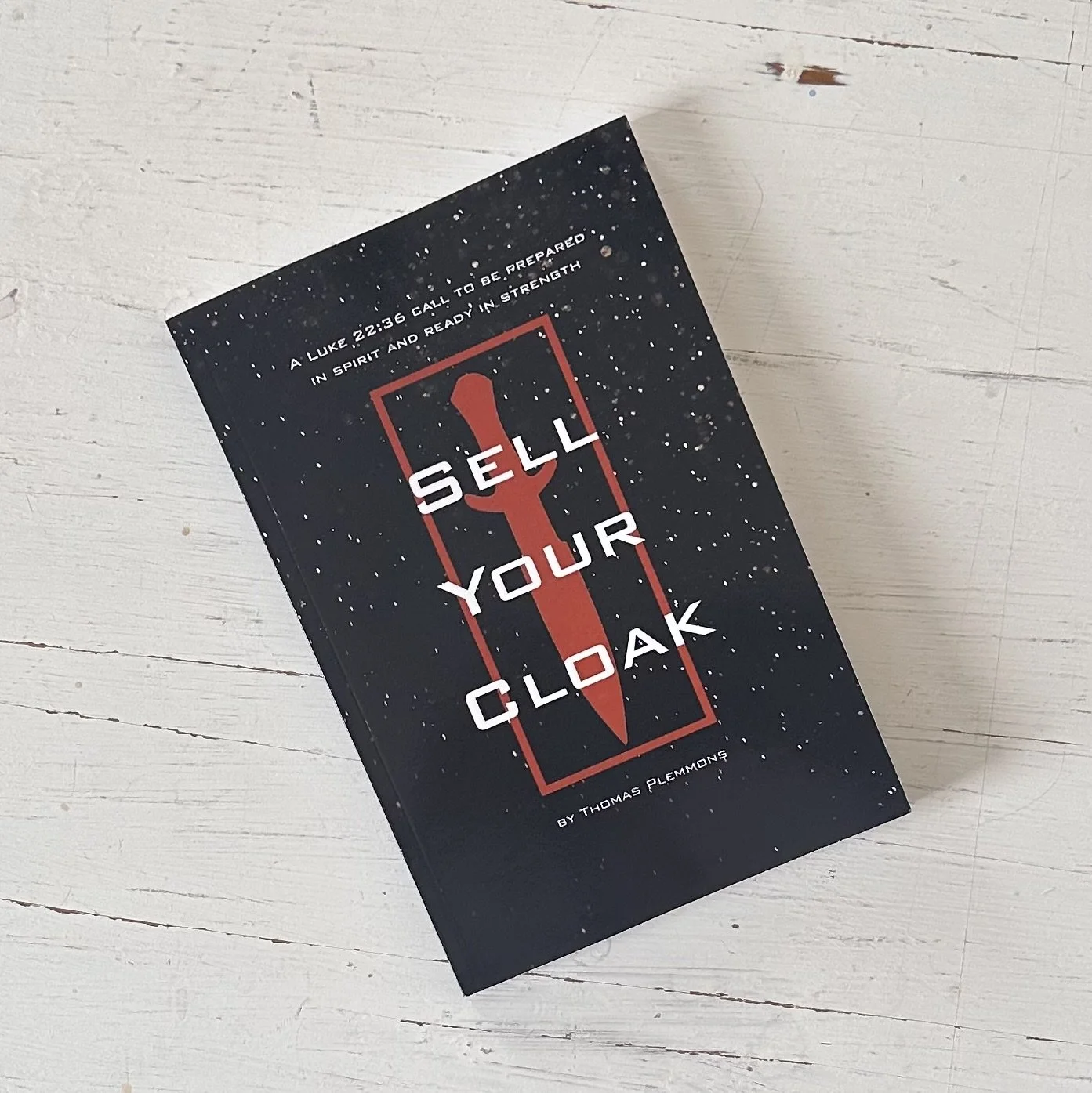 A book titled 'Sell Your Cloak' by Thomas Plemmons, featuring a black cover with white and red text, and a red dagger symbol, laying on a white wooden surface.
