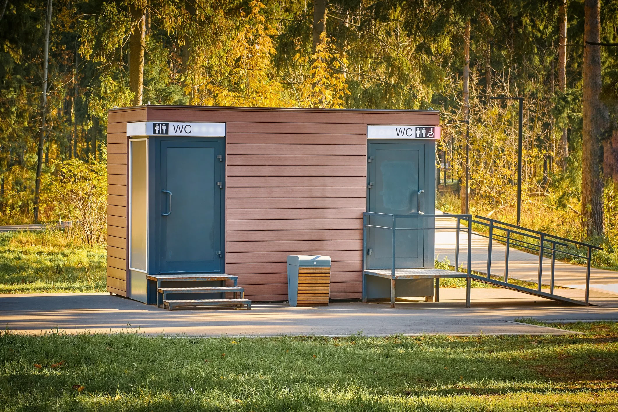 A small wooden building with two separate restroom doors, one for women and one for gender-neutral accessible use, situated in a park-like setting with trees and grass