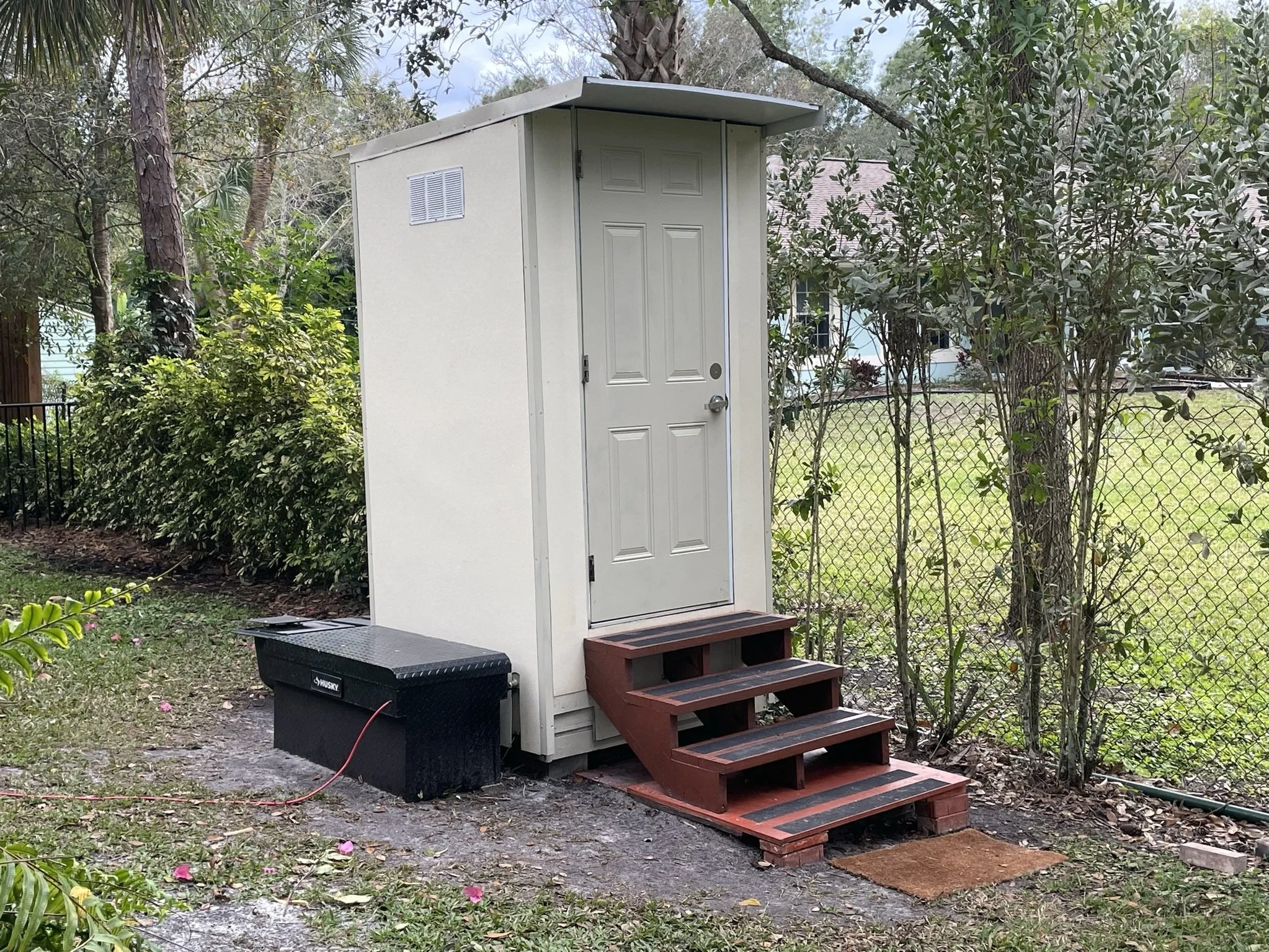 A small outdoor shed with a set of wooden steps leading to the door, situated next to a chain-link fence and surrounded by trees and bushes.