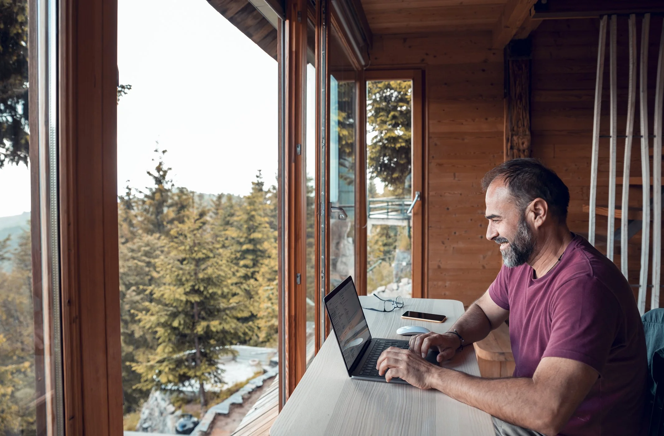 A man with a beard and short hair working on a laptop at a wooden desk in a room with large windows showing trees outside.