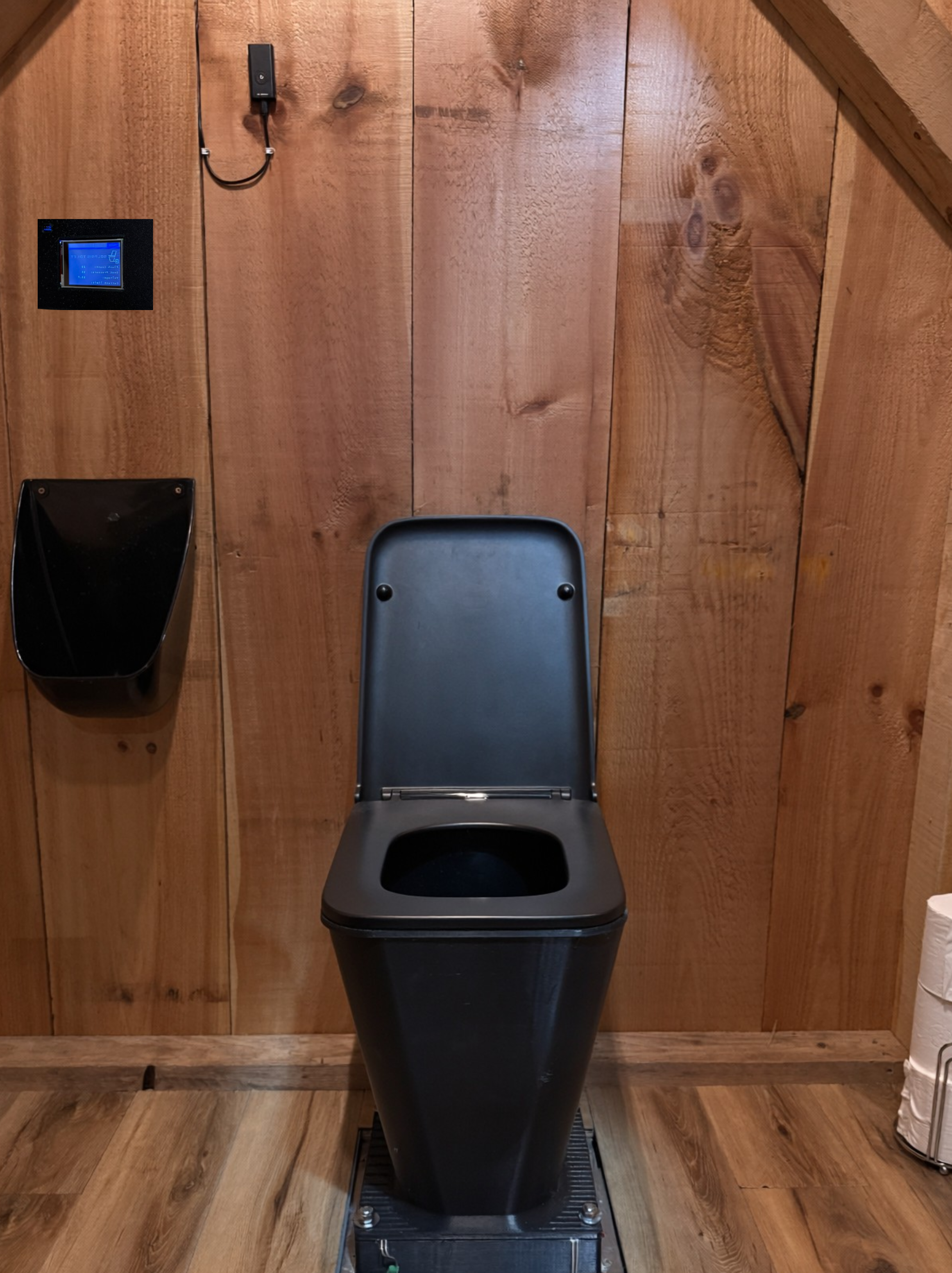 A black toilet with an open lid mounted on a wooden wall in a rustic bathroom. Above the trash bin is a small wall-mounted digital control panel. To the right of the toilet, part of a paper towel roll is visible on a metal holder.