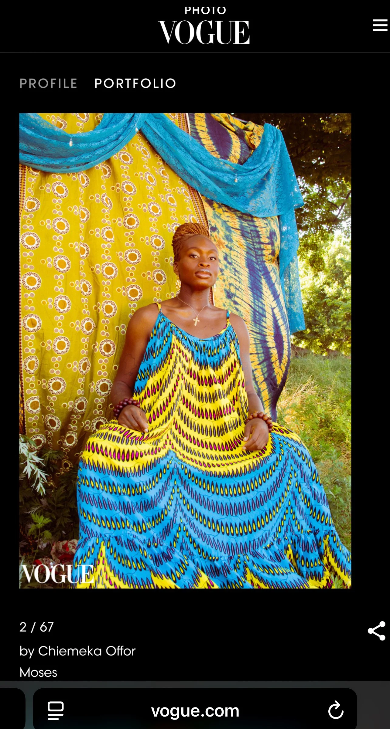 Moses Aina on Vogue. Com wearing a colorful patterned dress sitting outdoors in front of a vibrant yellow and blue fabric backdrop with floral designs and greenery.