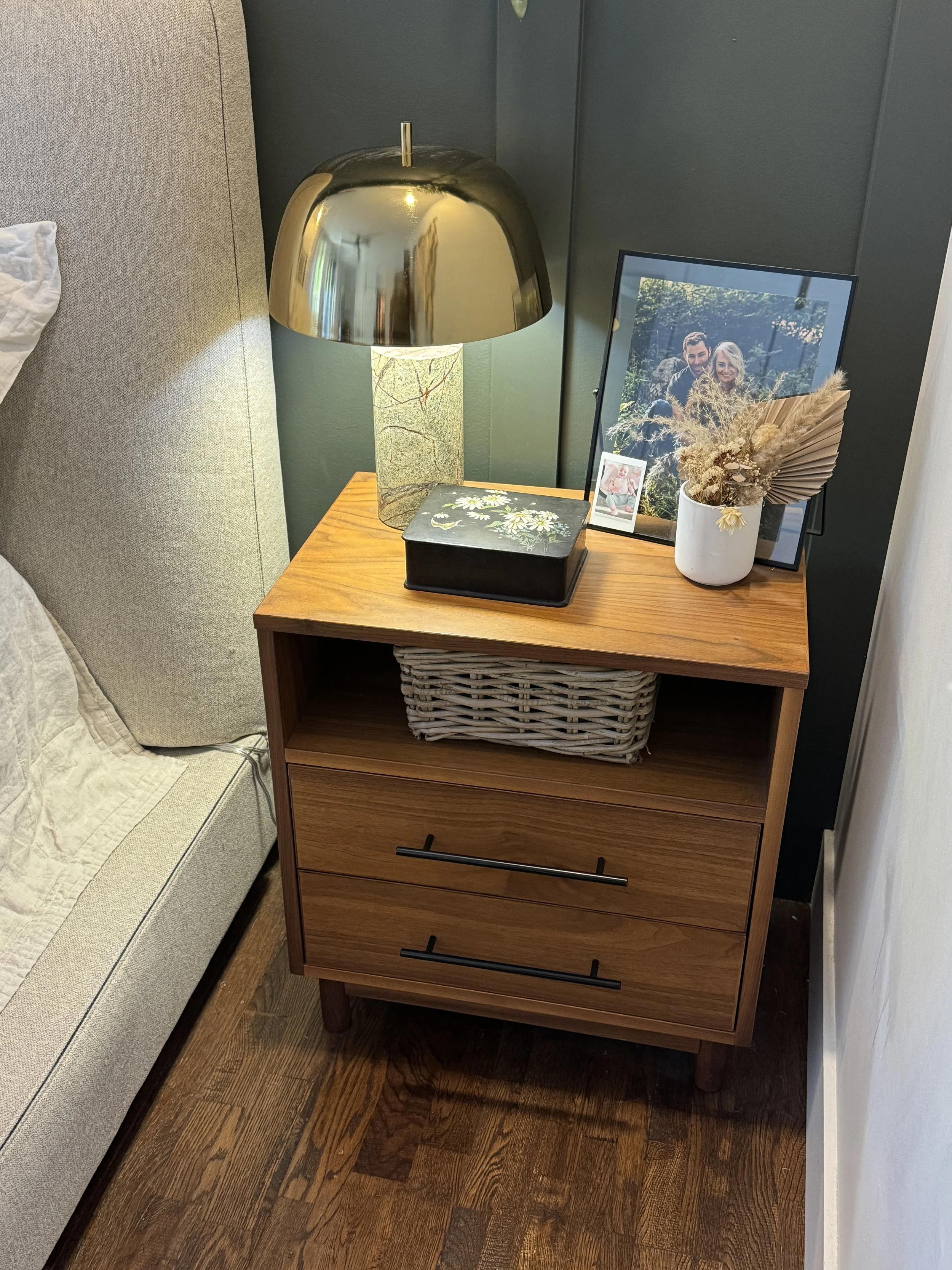 A wooden nightstand with two drawers and black handles, holding a glass table lamp with a gold shade, framed family photograph, small framed picture, and a white vase with dried flowers; a wicker basket is placed inside the open shelf of the nightstand.