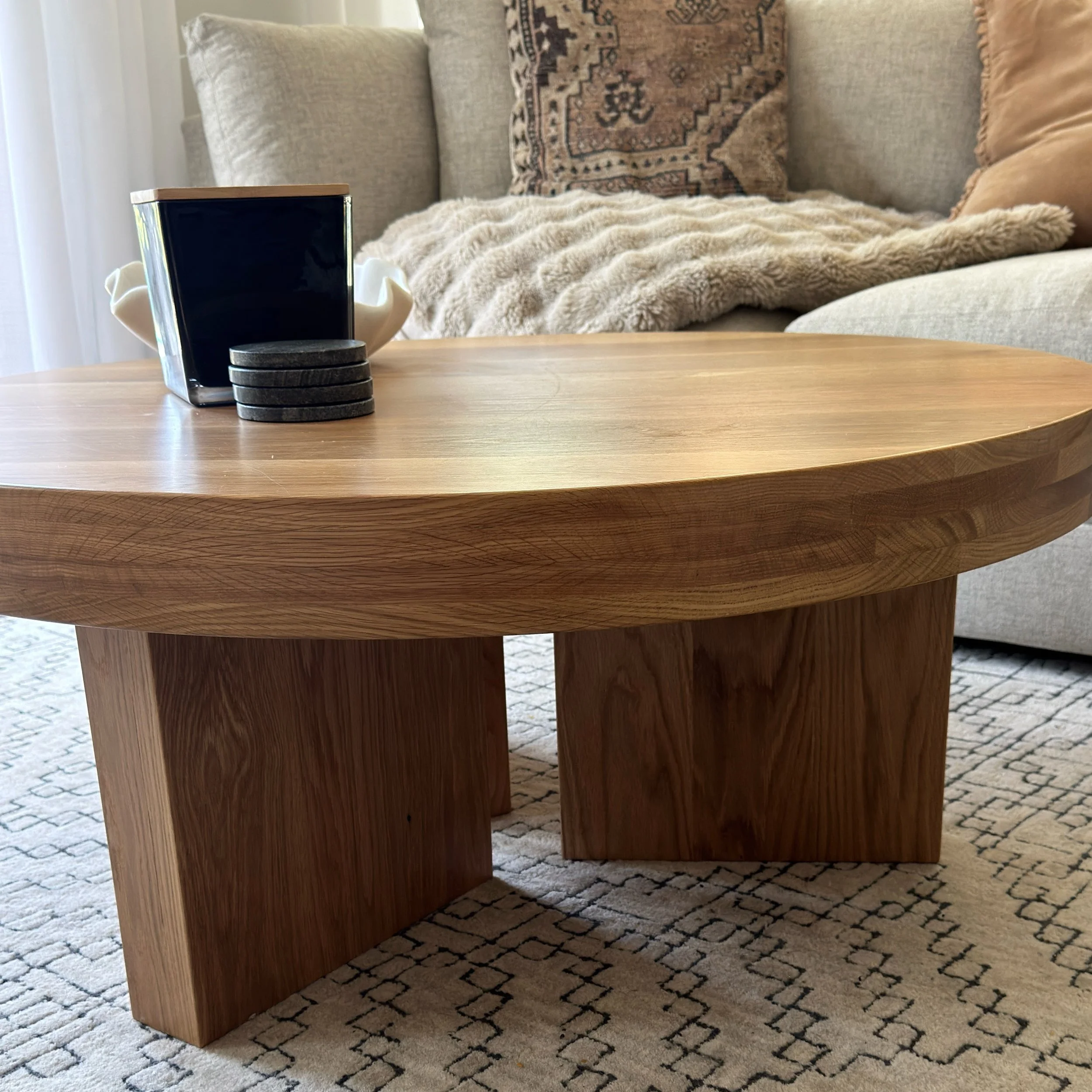 A round wooden coffee table with a black square-shaped item, four stacked round black coasters, and a white bowl on top, in a cozy living room with beige sofa and cushions, a patterned throw, and a beige rug.