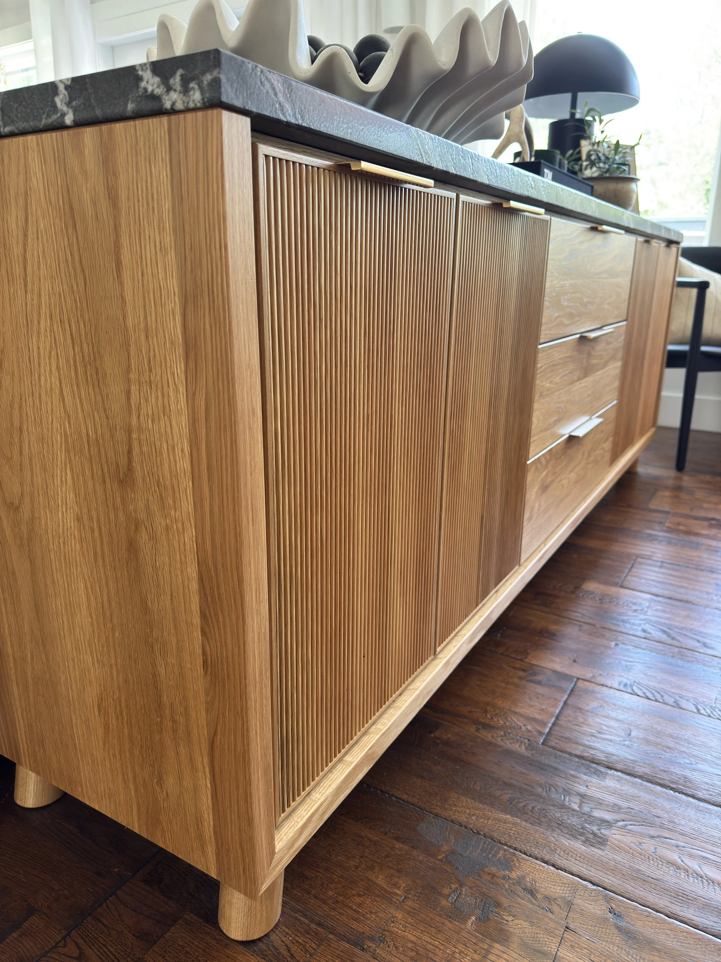 Close-up of a mid-century modern wooden sideboard with textured vertical slats on the doors, set on a hardwood floor with a dark marble top surface, decorated with ceramics and a modern black lamp in the background.