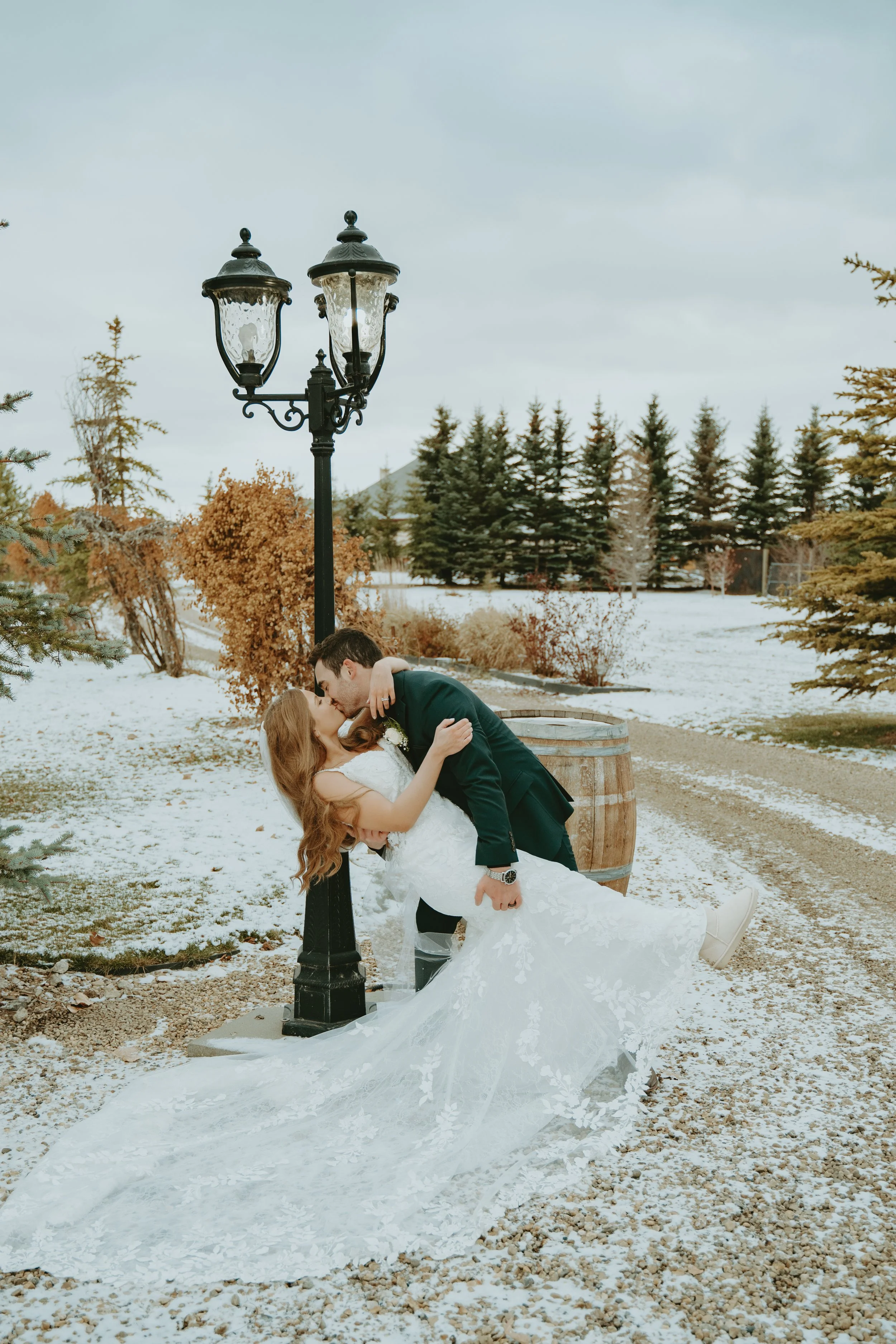 A bride and groom share a kiss outdoors on a snowy day, with the groom dipping the bride and a lamppost nearby.