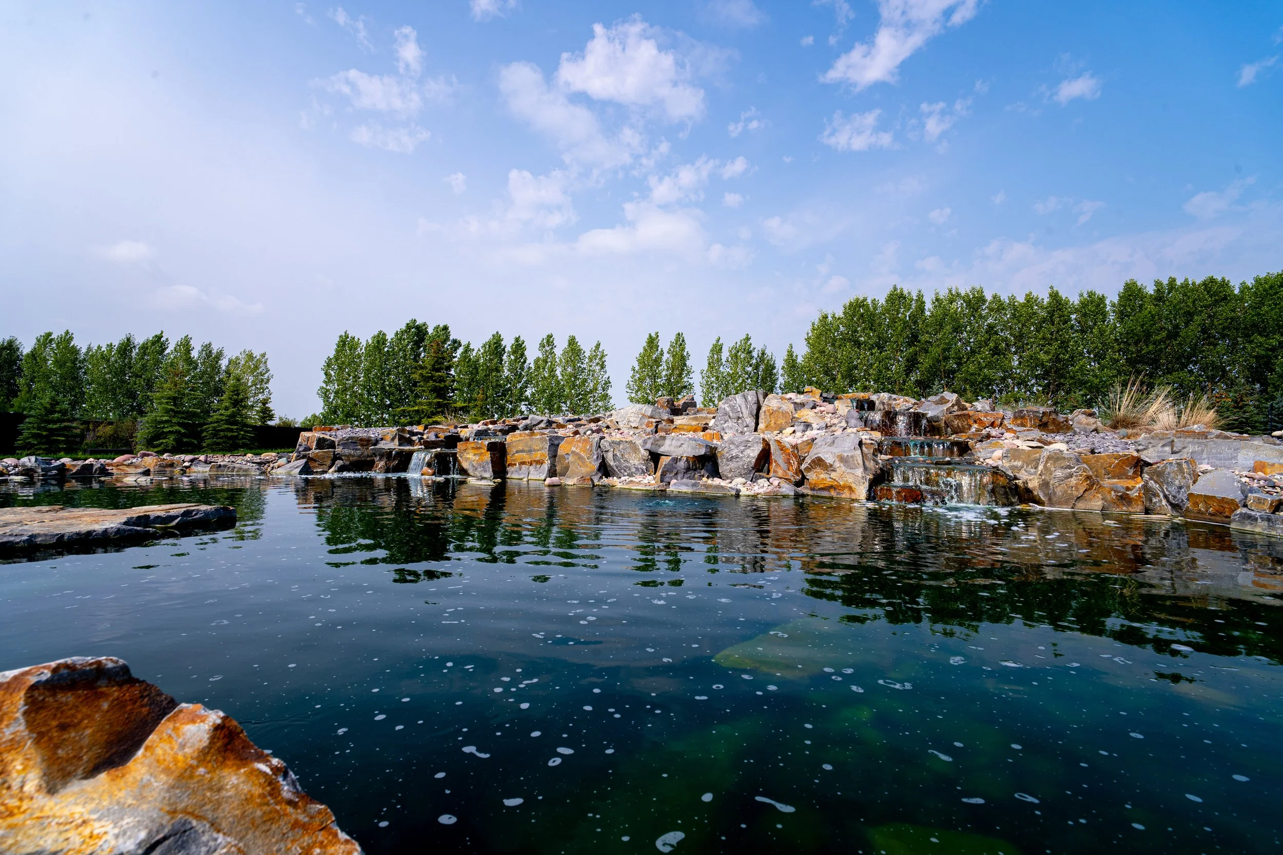 A pond with rocks and small waterfalls, surrounded by green trees under a partly cloudy sky.