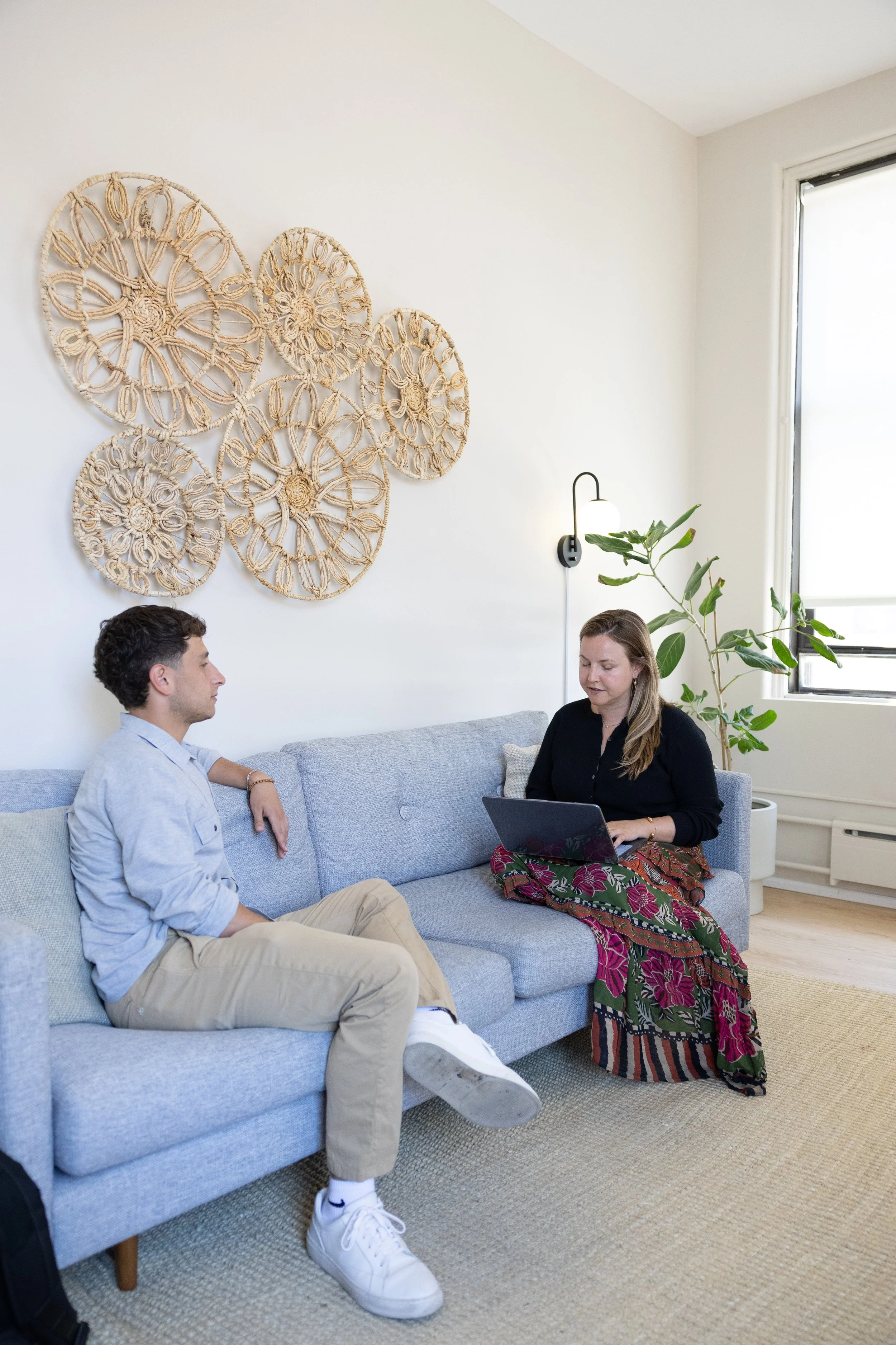 Two people sitting on a gray sofa in a bright room. The man on the left is wearing a light blue shirt and khaki pants, while the woman on the right has a laptop on her lap and is wearing a black top and a colorful skirt with a floral pattern. Behind them, there is wall decor made of woven circular patterns, a black wall sconce with a white light bulb, a large green potted plant, and a window with sunlight coming through.