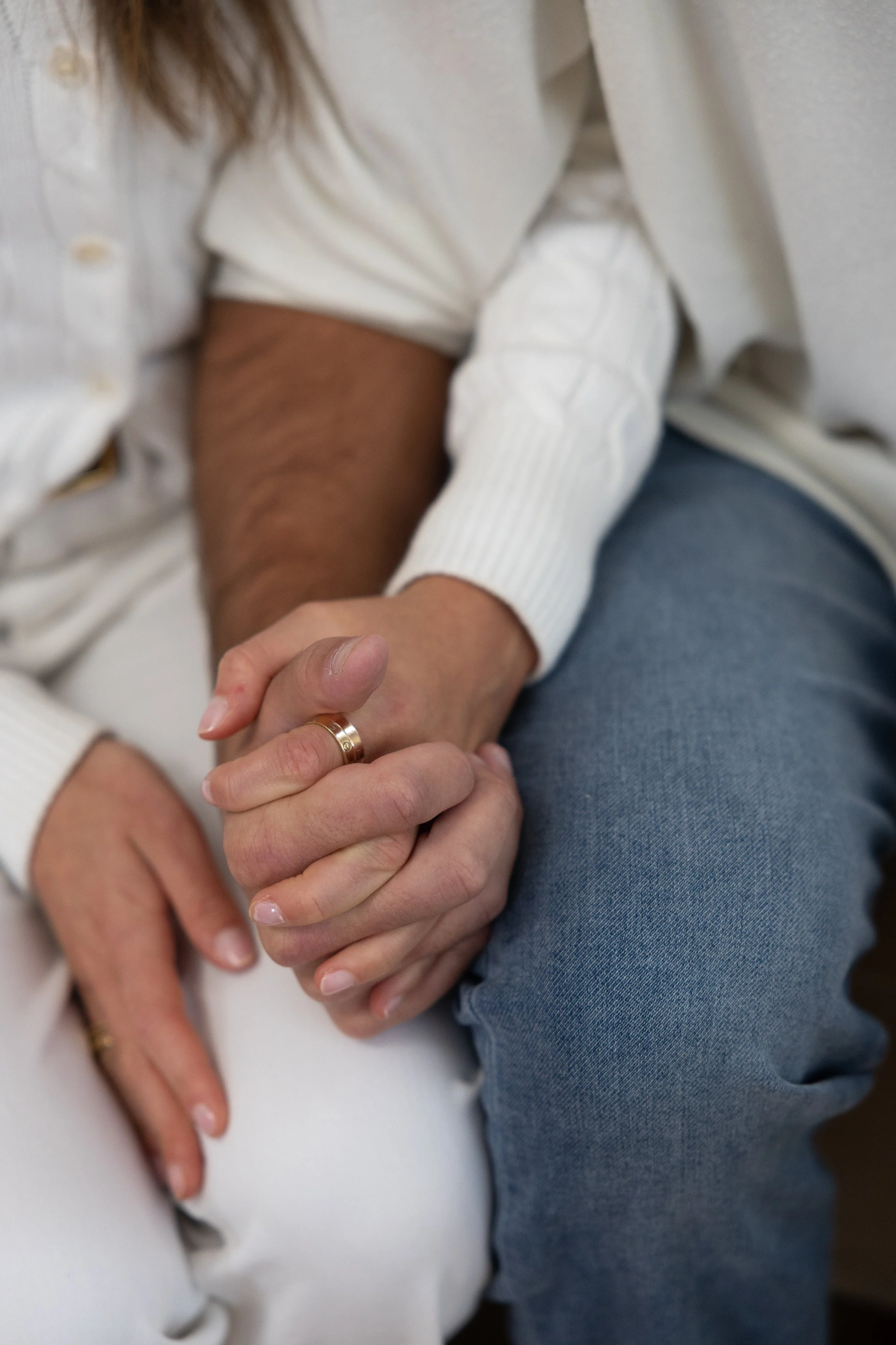 Two people holding hands, one with a wedding band, sitting close together. The person's arm is resting on their lap, and they are wearing a white sweater, while the other individual is wearing a light-colored shirt and jeans.