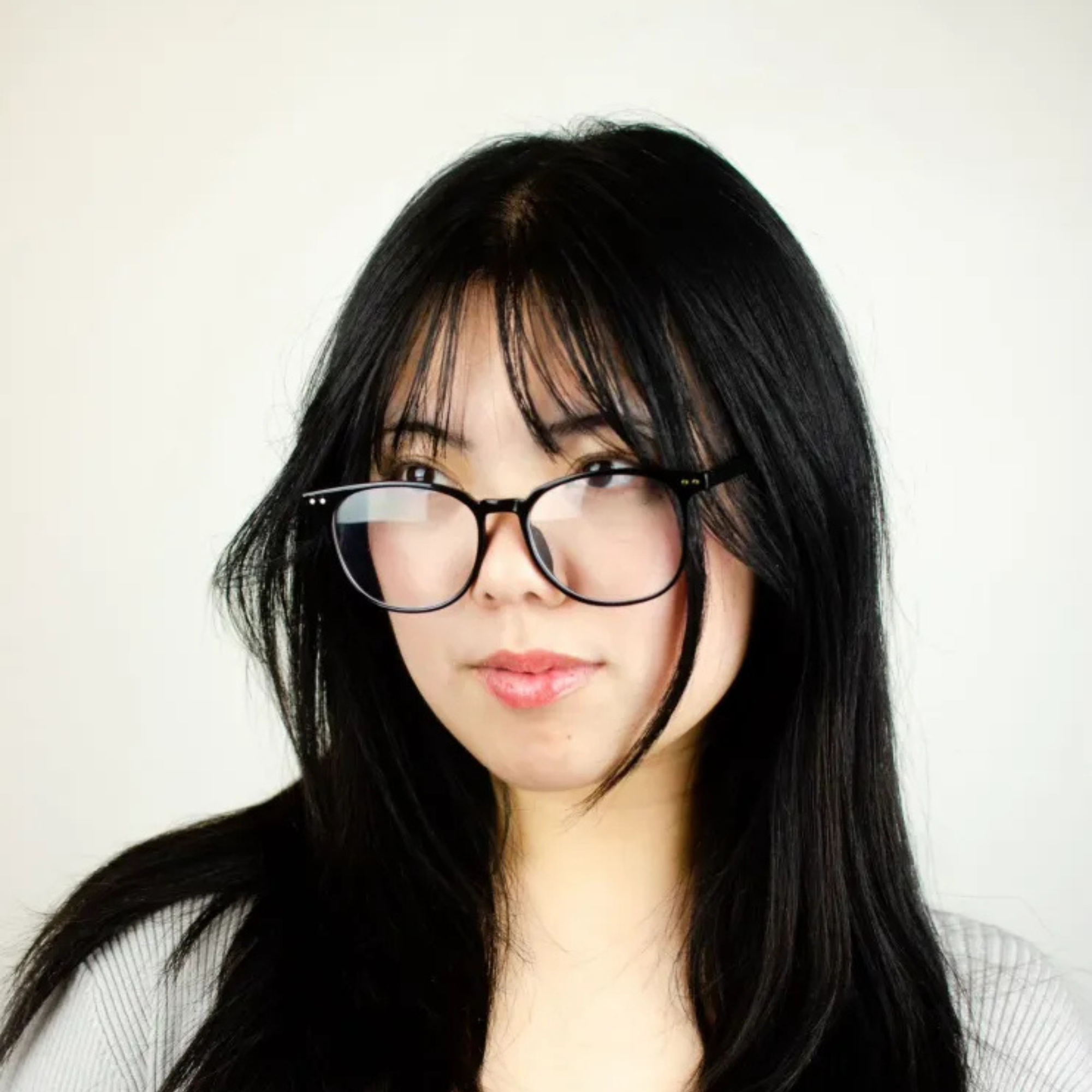 A woman with long black hair and glasses, looking slightly to the side with a neutral expression, in front of a plain light-colored background.