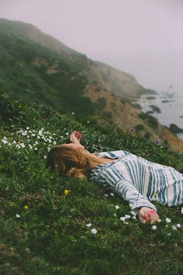 A woman lying on a grassy hillside surrounded by small white and yellow flowers, with cliffs and the ocean in the background on a cloudy day.