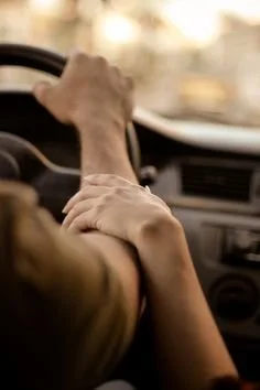 A person driving a car, holding the steering wheel with one hand while the other hand rests on their arm.