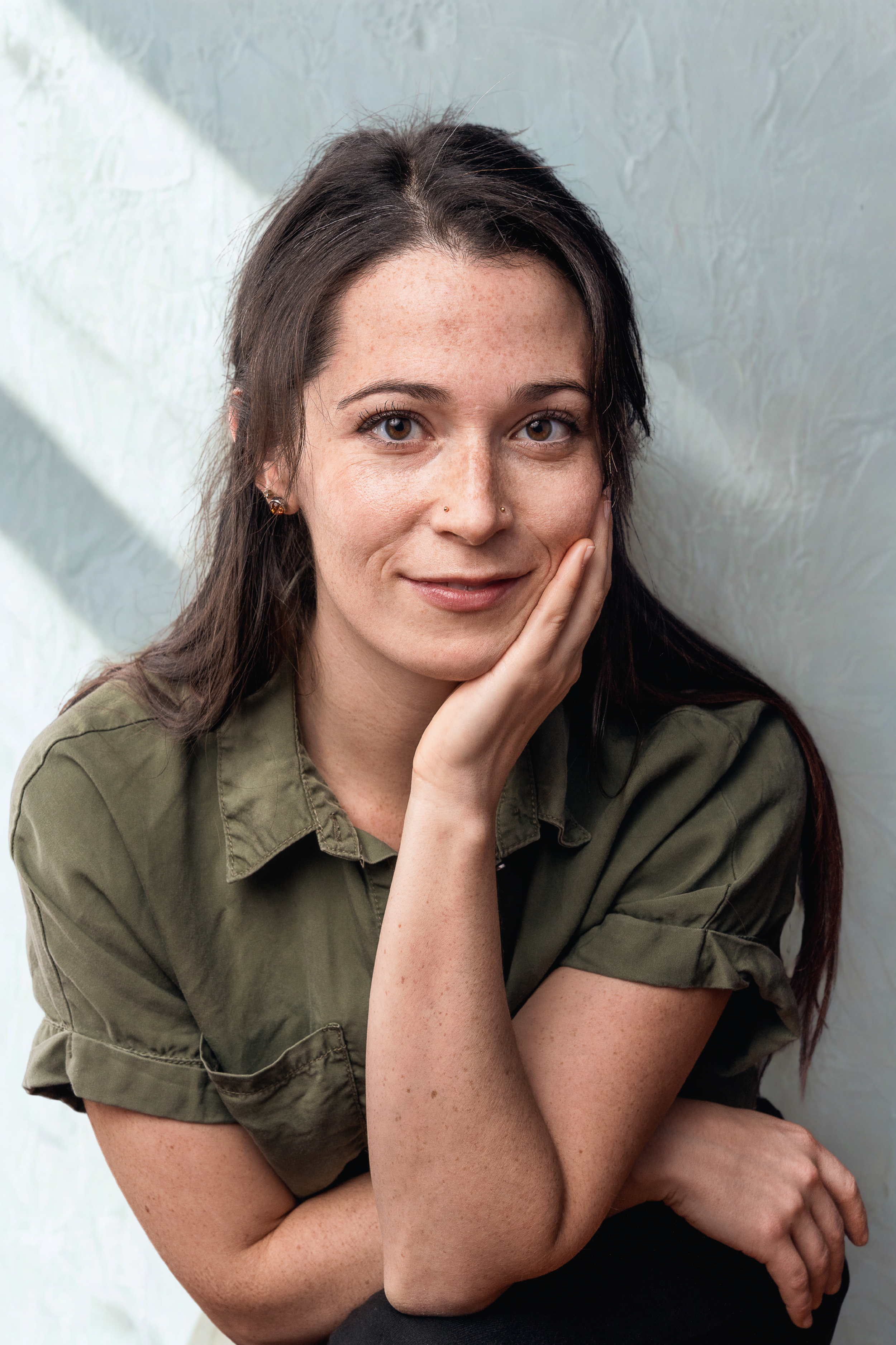 A woman with long dark hair and light skin, wearing a green shirt, smiling and resting her face on her hand, sitting in front of a textured light-colored wall.
