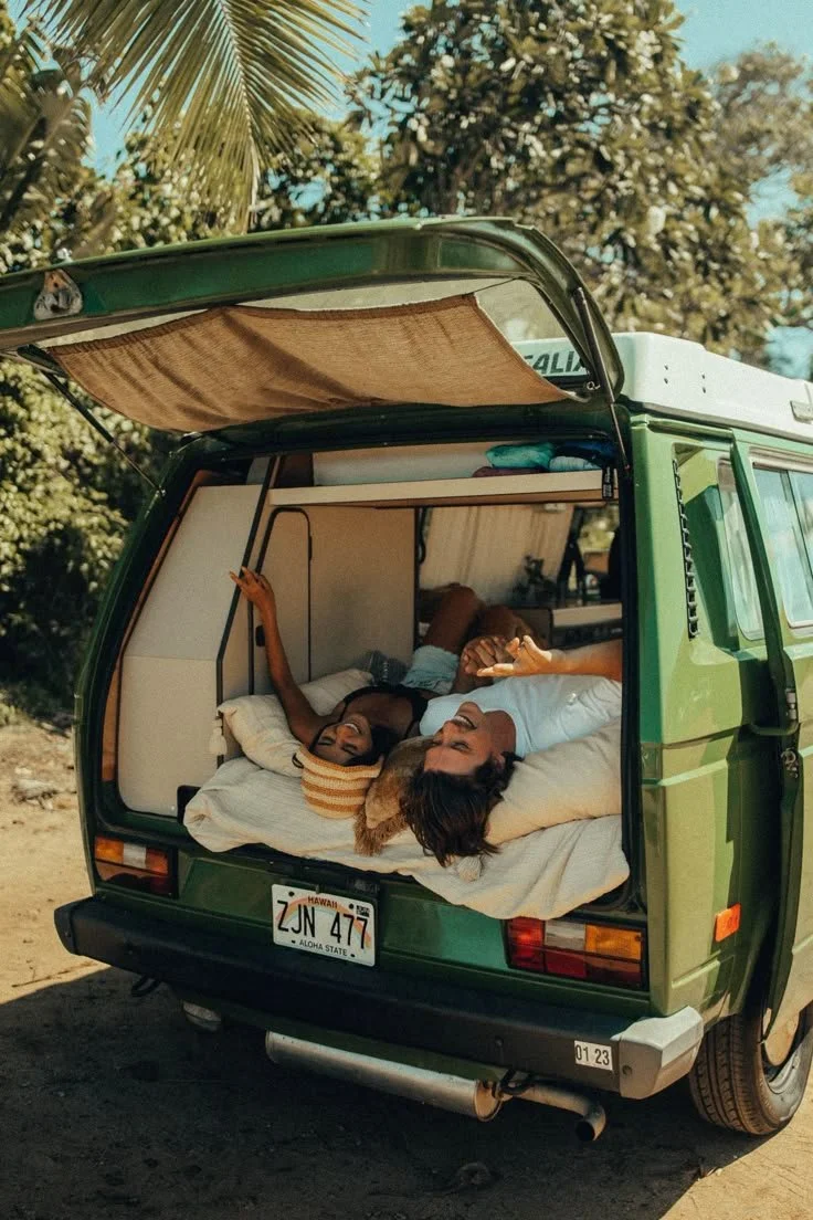 Two women are lying inside the open back of a green camper van, relaxing and smiling in a sunny outdoor setting surrounded by trees.