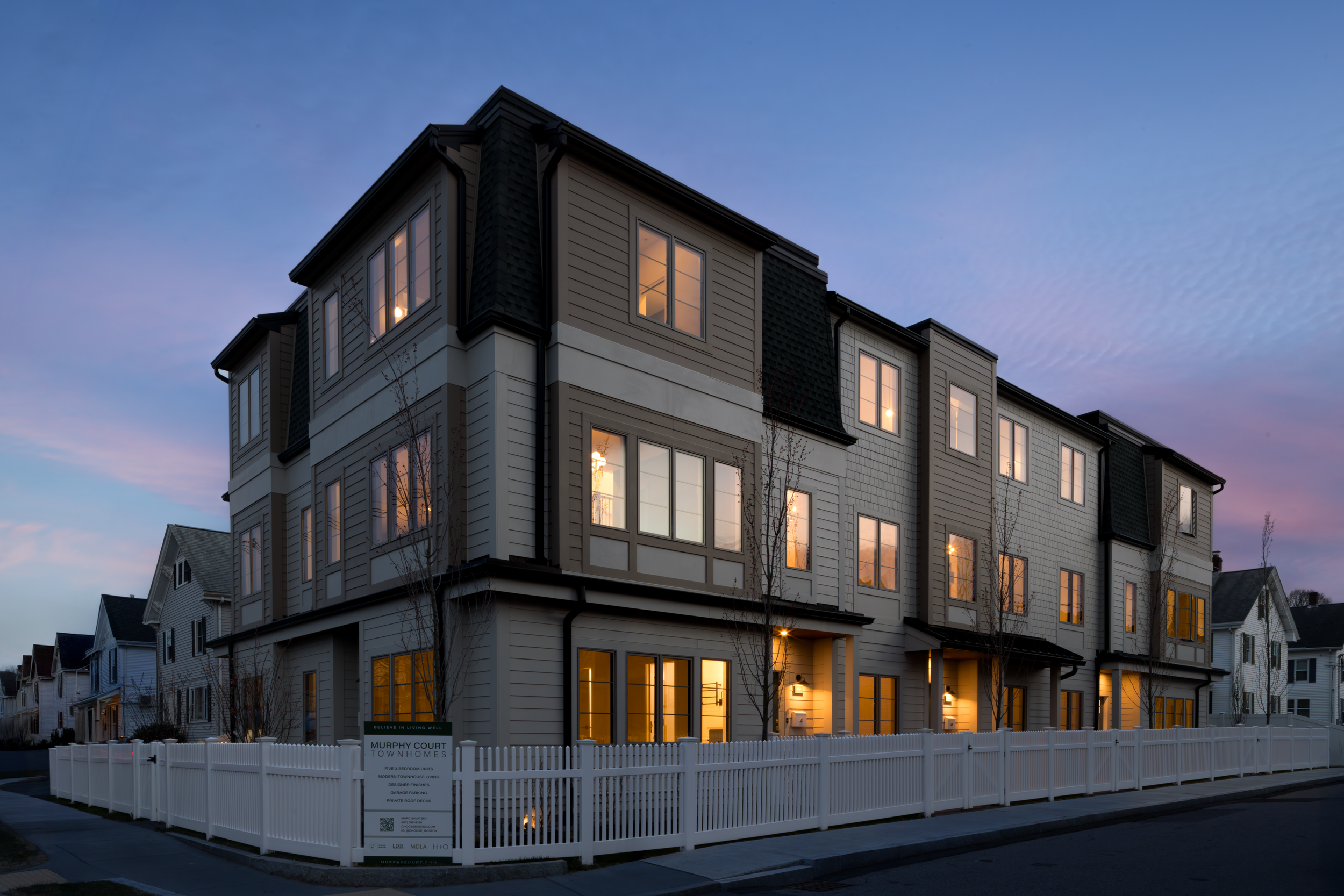 A multi-story residential building with large windows, illuminated from within, surrounded by a white picket fence and a sidewalk, during evening twilight.