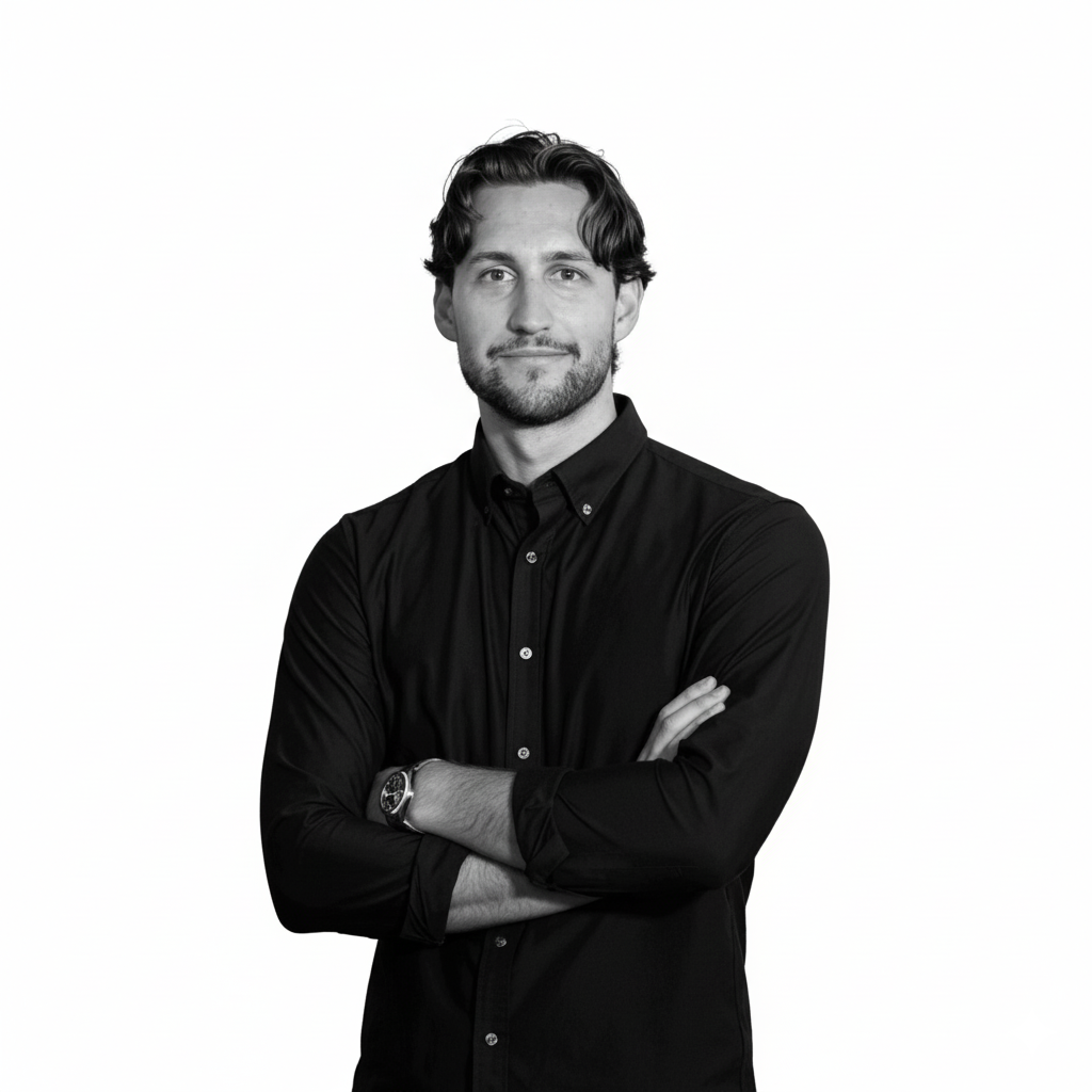 Black and white portrait of a young man with dark hair and a beard, wearing a dark button-up shirt with folded sleeves, crossing his arms, against a plain white background.