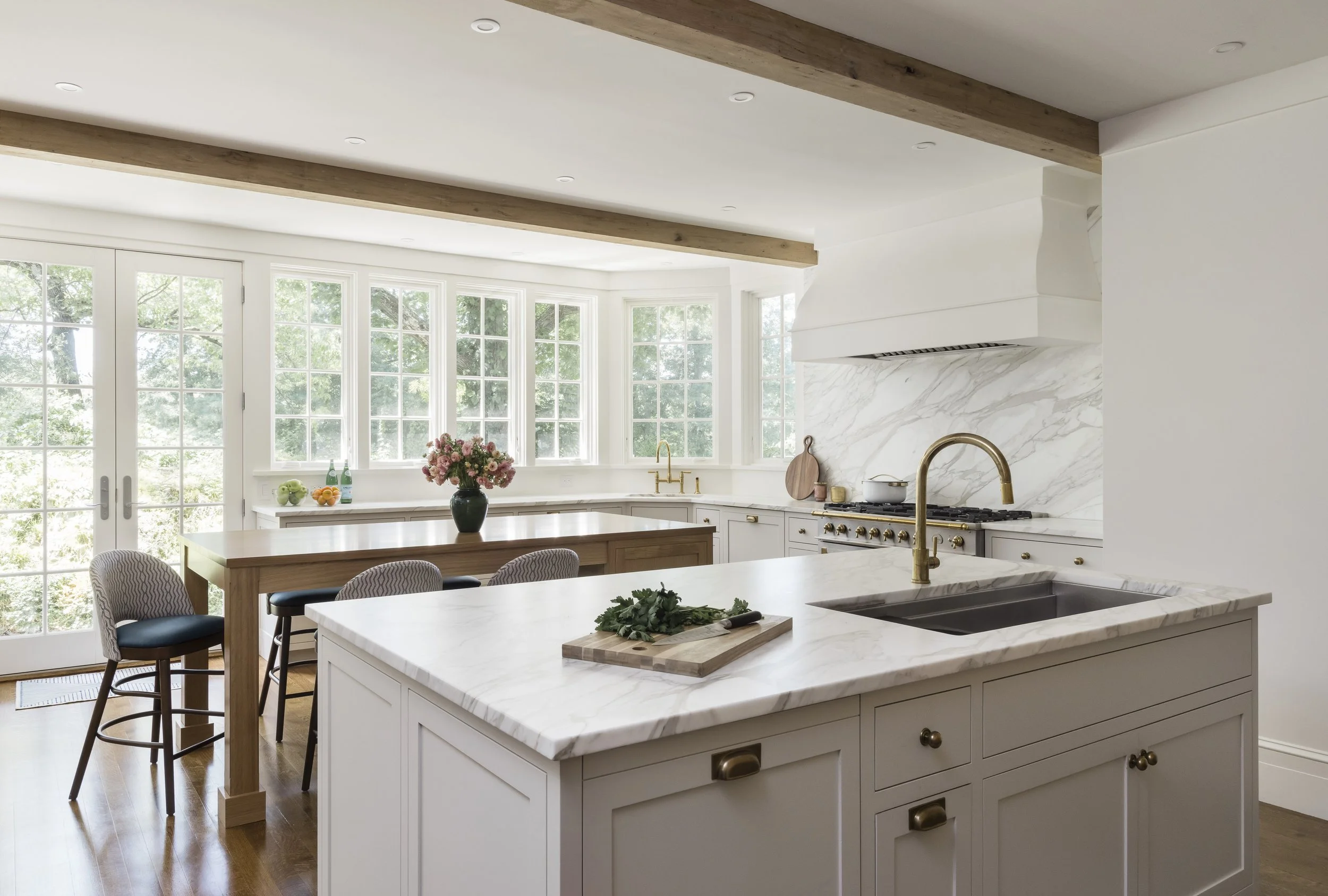 A modern kitchen with white cabinetry and marble countertops. There is a large kitchen island with a wooden table and chairs nearby. The back wall has tall windows letting in natural light, and there are decorative flowers and bottles on the window s