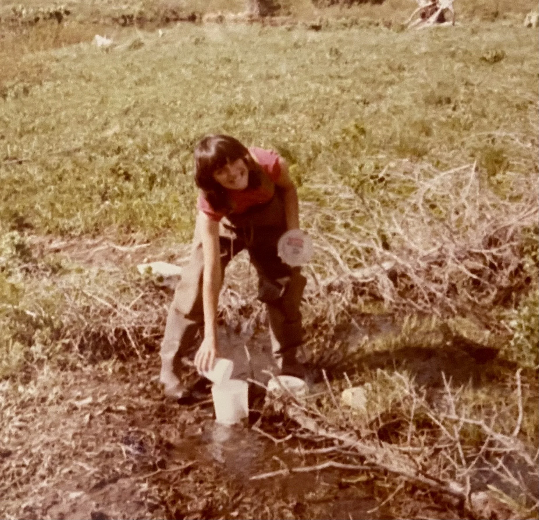 A young girl harvesting water from a small stream into white buckets in a rural outdoor area.