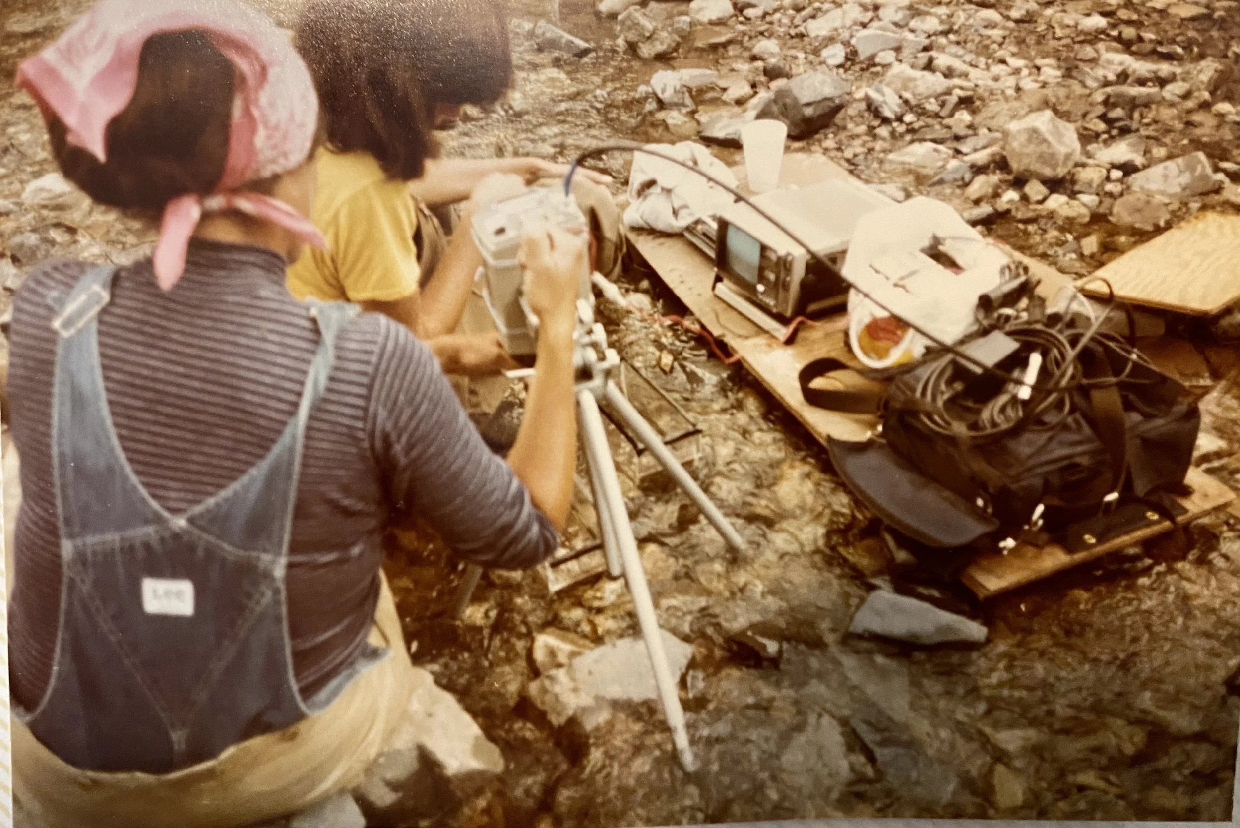 Two women working with geological equipment on rocky terrain, surrounded by various tools, cables, and a cup.