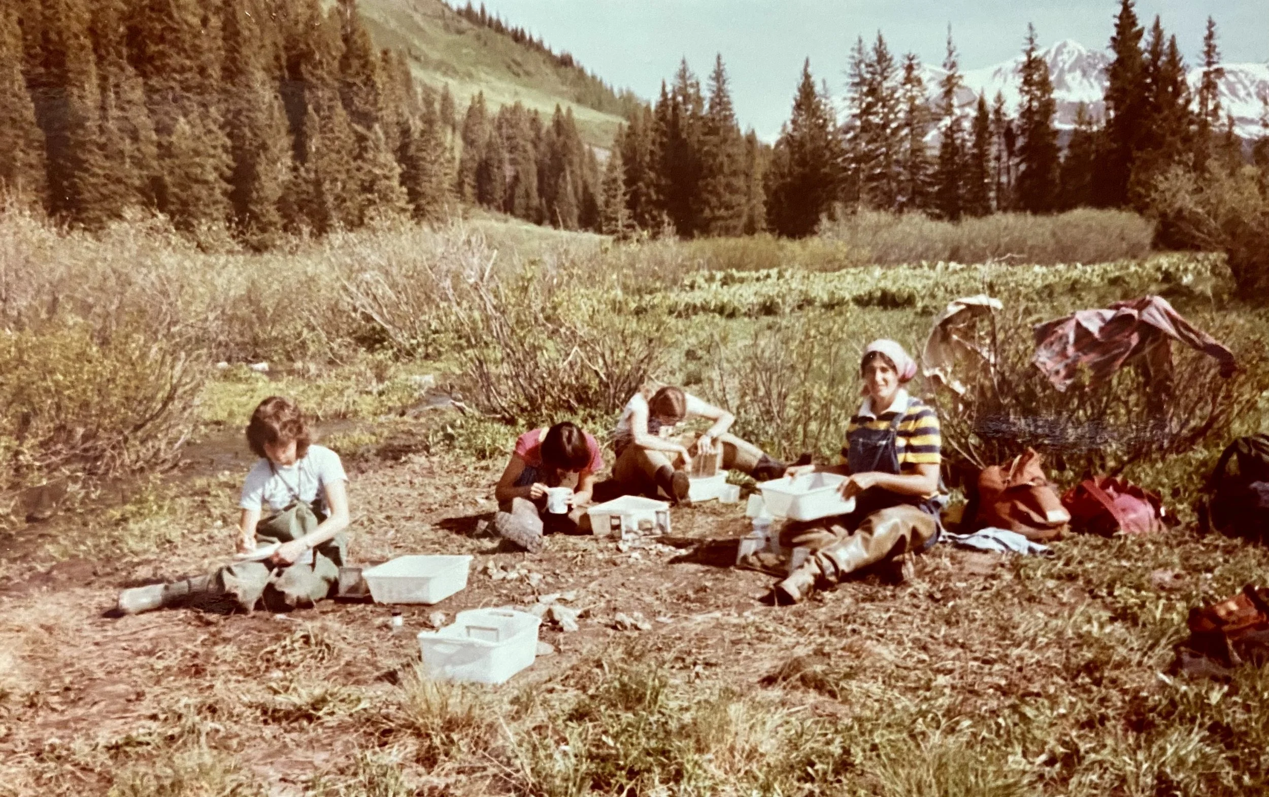 Four people, including children, are sitting on the ground outdoors in a mountainous forested area, eating together with food containers and backpacks around them.