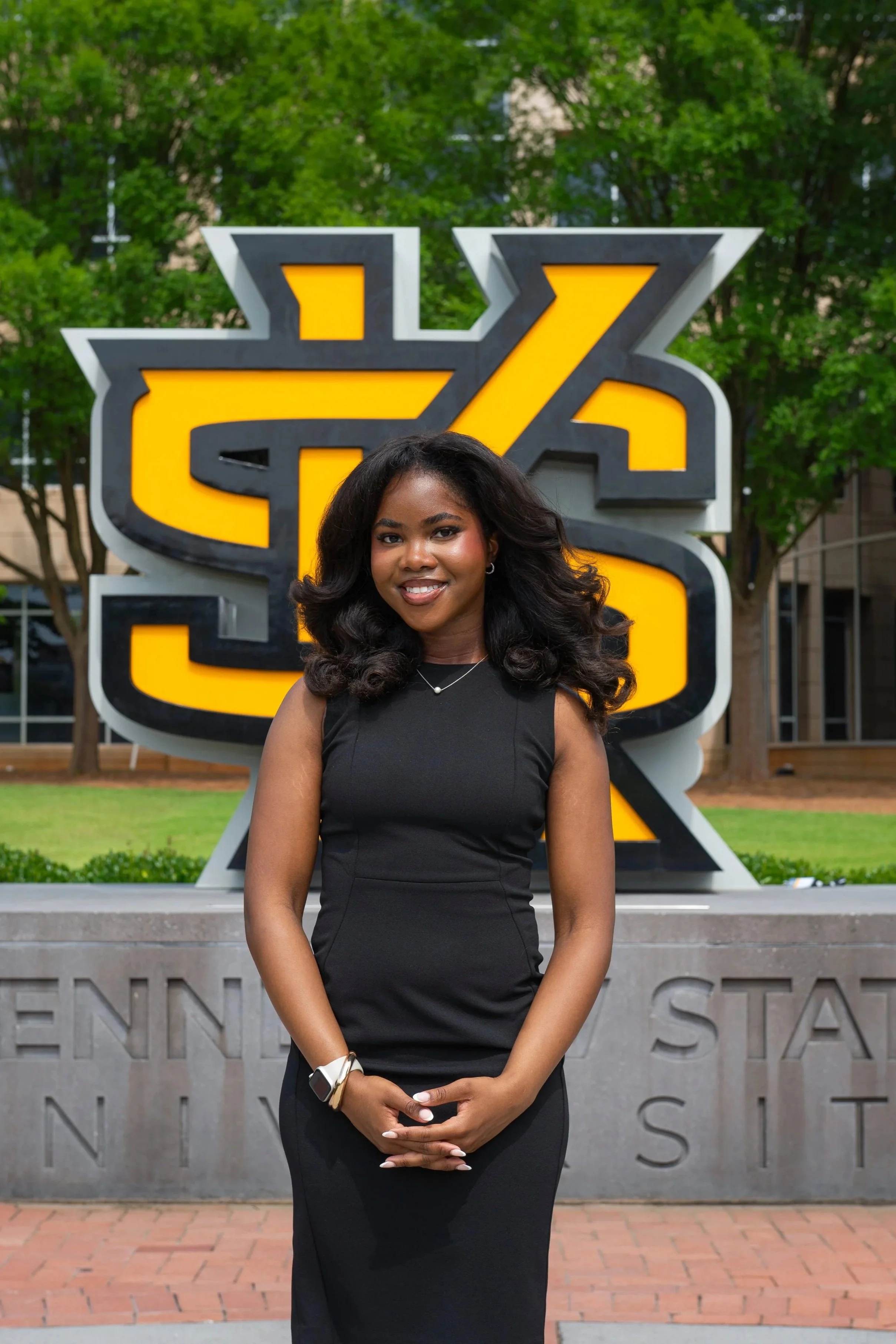 A smiling woman in a black blazer and skirt standing outdoors at a university sign.