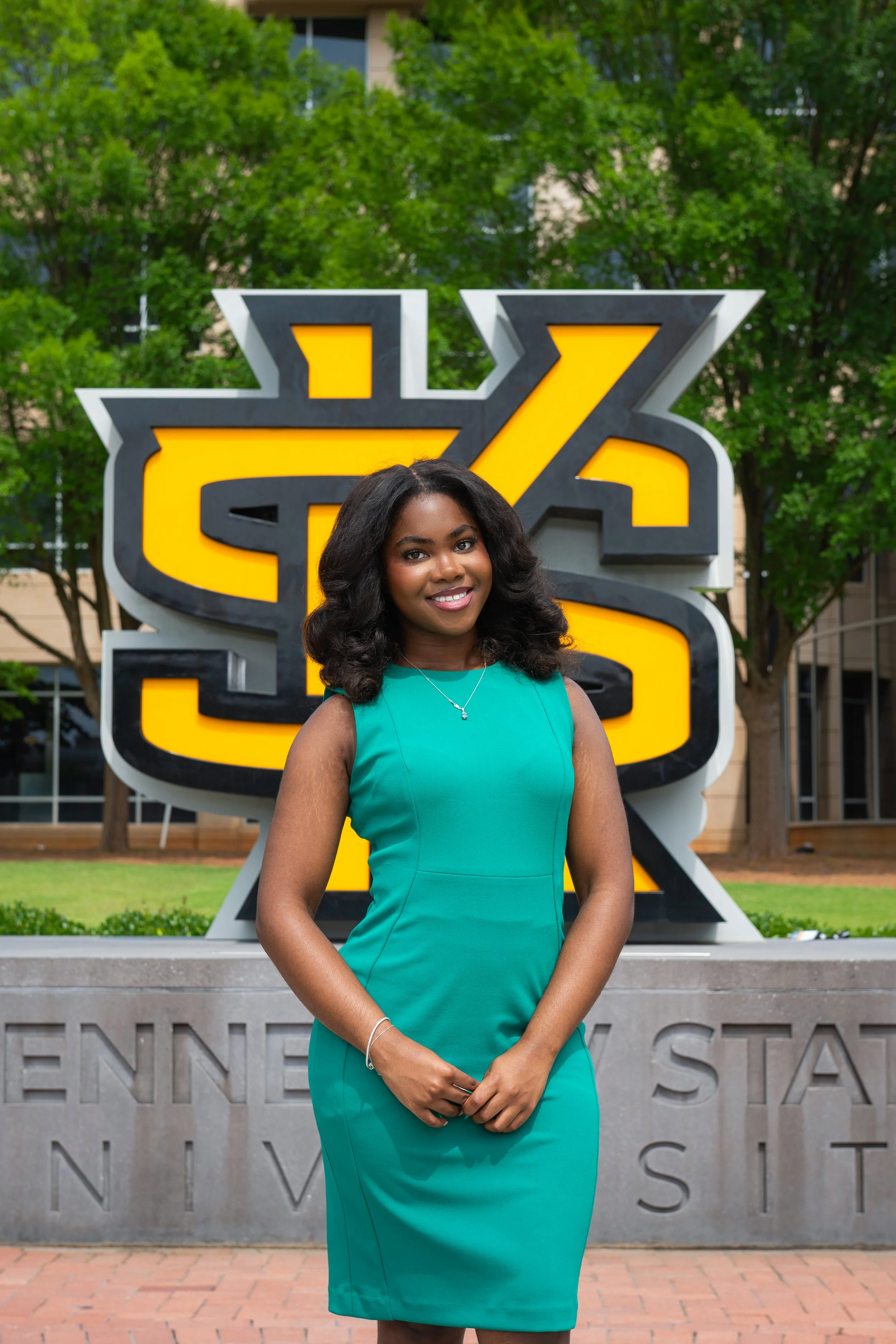 A woman standing outdoors in front of a large decorative sign featuring a stylized yellow and black design. She has curly hair, is dressed in a dark blazer and a teal blouse, and is smiling slightly.
